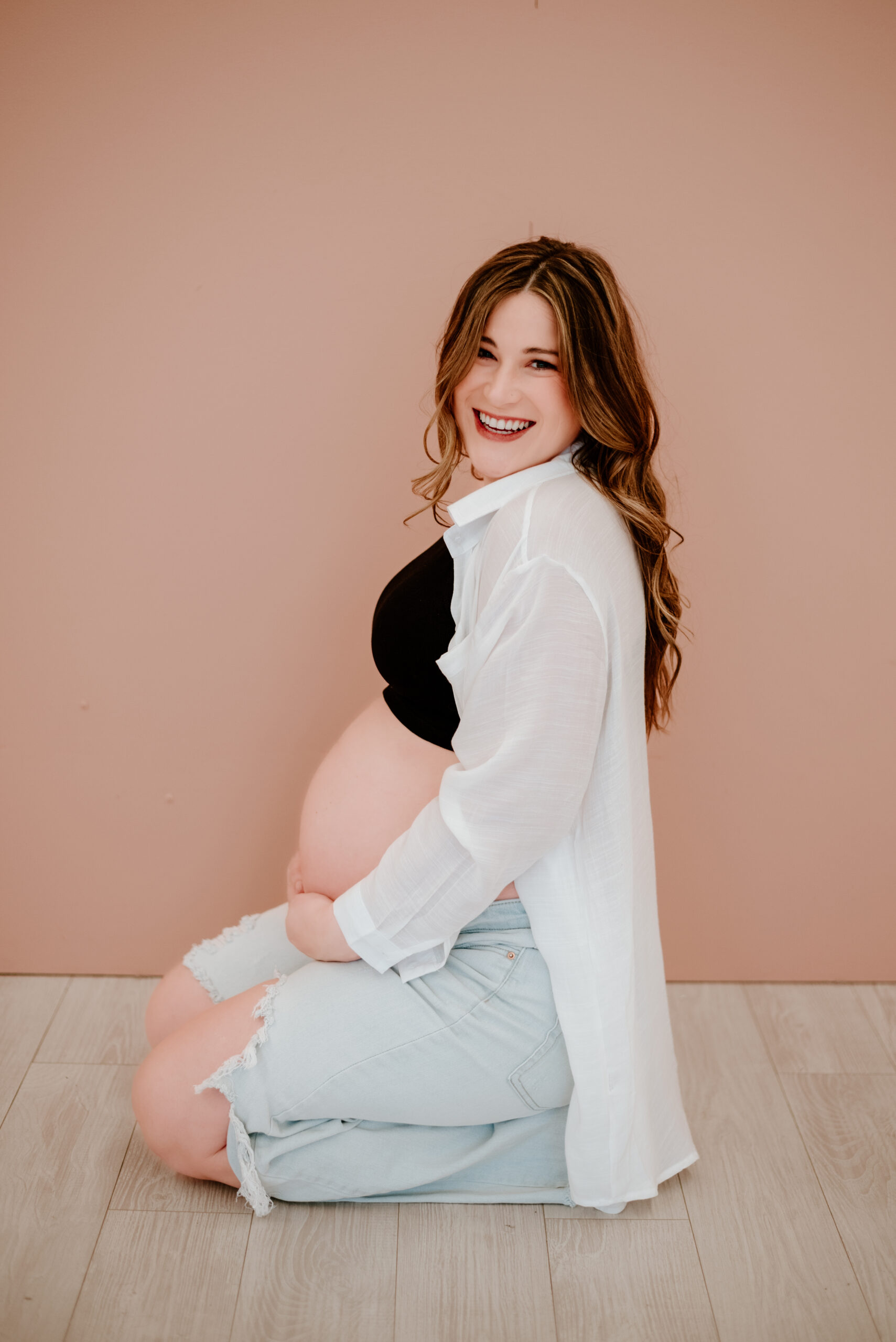 a pregnant woman poses in front of a pink wall on her knees