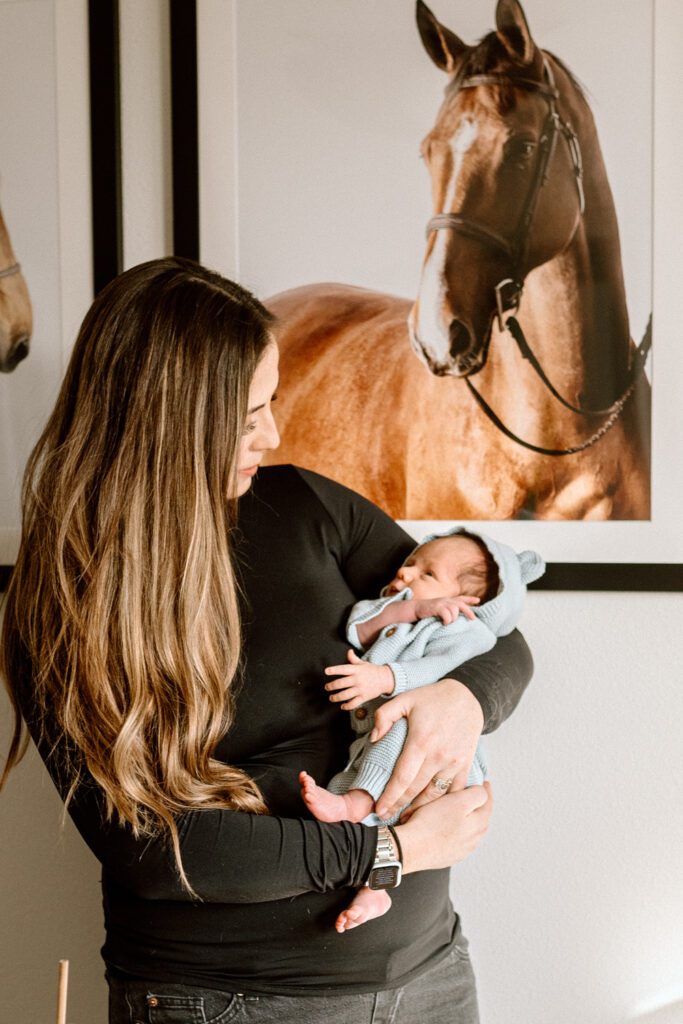 a mom holds her newborn baby in front of a framed photo of a horse