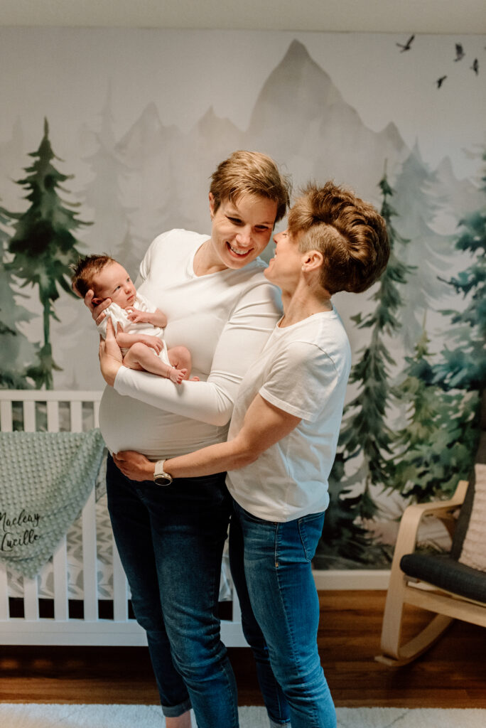 two moms stand in front of the crib in a nursery holding their newborn baby