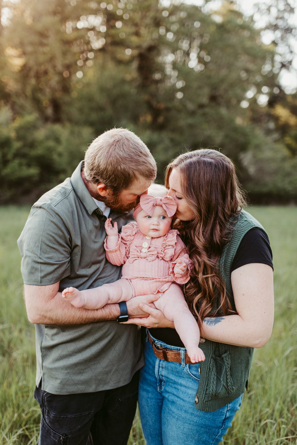 a mom and dad hold thier baby between them each kissing a cheek in an open field
