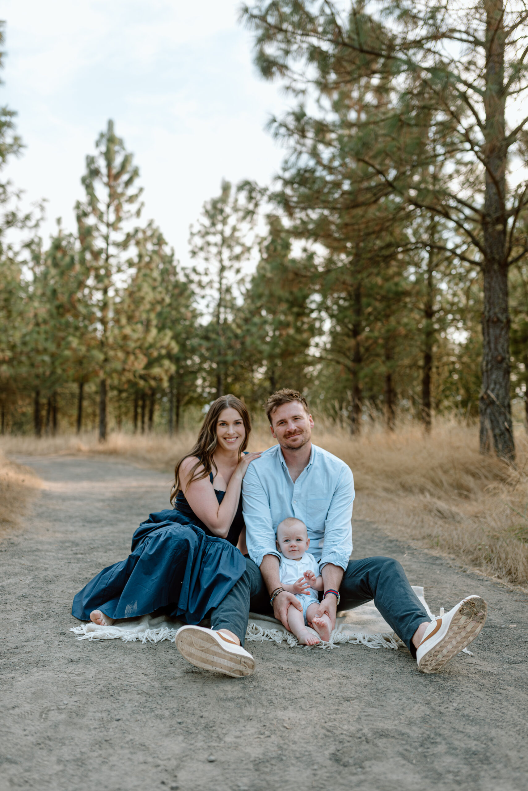 mom, dad, and baby sit on a blanket in the middle of a gravel path at a nature park